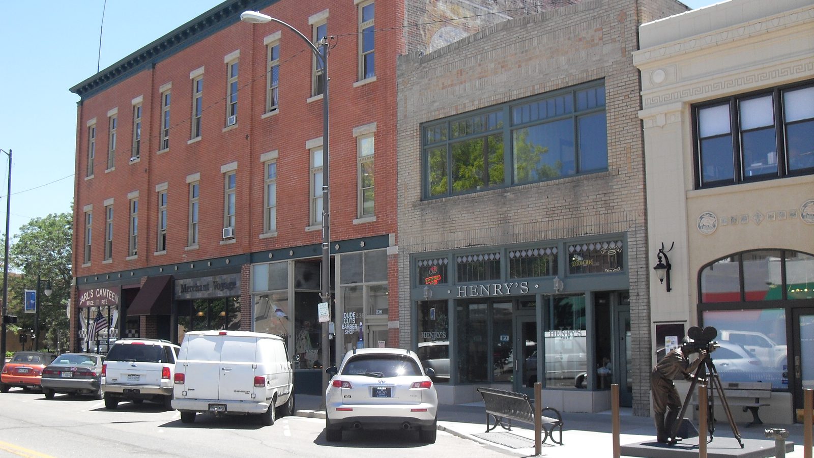 Two-storey historic brick commercial buildings along 4th Street in downtown Loveland, with parked cars and a bronze sculpture of a cameraman on the sidewalk